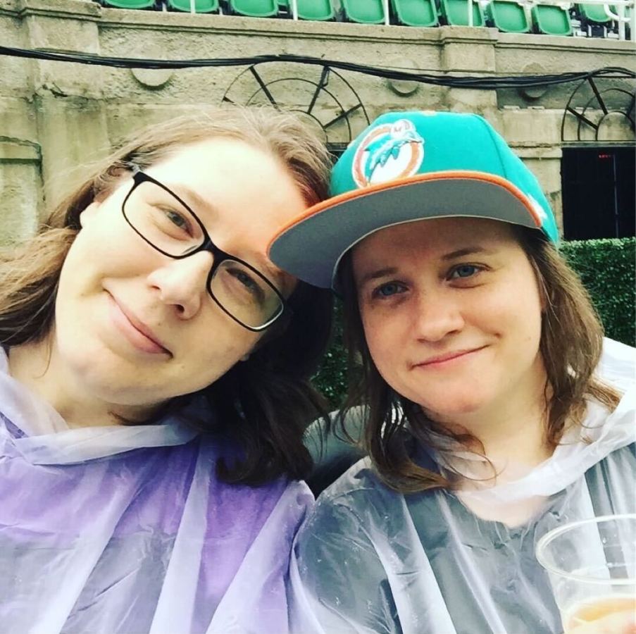 Heather and Stacy are both wearing rain ponchos in this photo wear they are also both smiling. They are both white women with medium length brown hair. Heather has black framed glasses and Stacy is wearing a baseball cap.