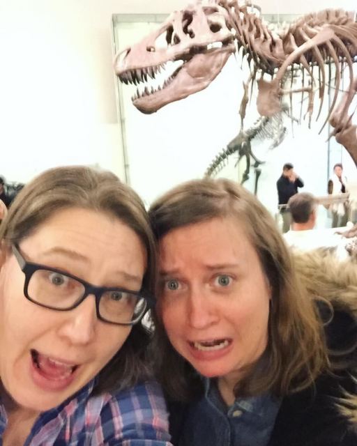 Heather, a soft butch white woman here with brown hair and dark framed glasses gasps in mock-fear while Stacy a white woman with medium length brown hair similarly gasps in mock fear, both in front of a museum T Rex skeleton