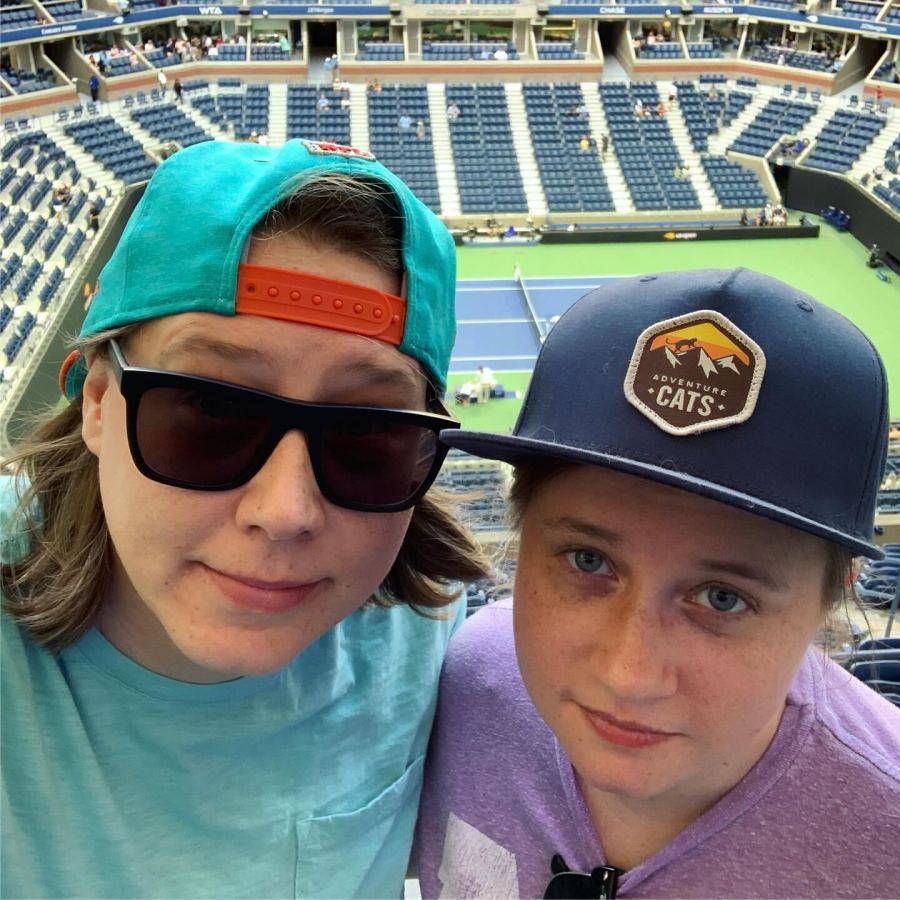 Heather and Stacy pose together at a Tennis game in a stadium. Heather is wearing a backwards baseball cap and sunglasses. Stacy is wearing an Adventure Cats baseball cap and lavender shirt. Both are white women with light brow hair.