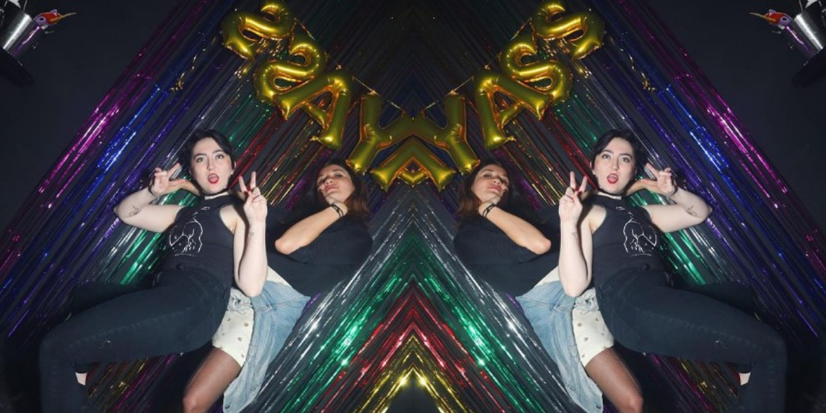 This feature image is made up of one image that is flipped and doubled on the other side, creating a mirror effect. The photo is of Analyssa and Courtney at Gay Asstrology. Analyssa is a a young latinx person with shoulder length brown hair, wearing shorts and a navy shirt with a jean jacket around her waist. She is leaning back against a bright and colorful background of rainbow streamers. Courtney is a white woman, and in front with short black hair, wearing all black with red lipstick, and giving double V for Victory signs with her fingers.
