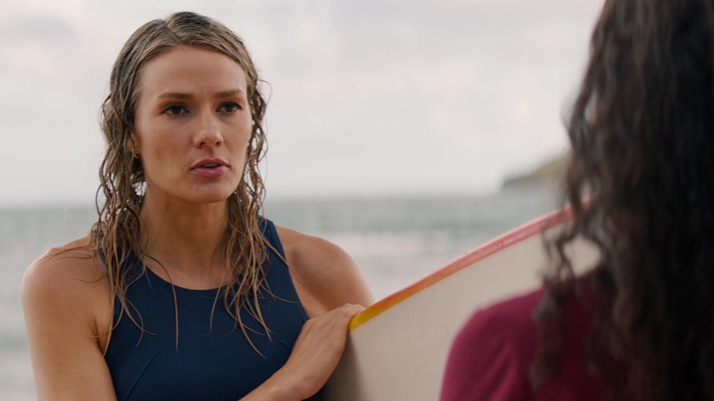 Kate approaches Lucy after she gets out of the ocean following a morning of surfing. She's in a bathing suit and carrying a white surfboard with red and yellow trim.