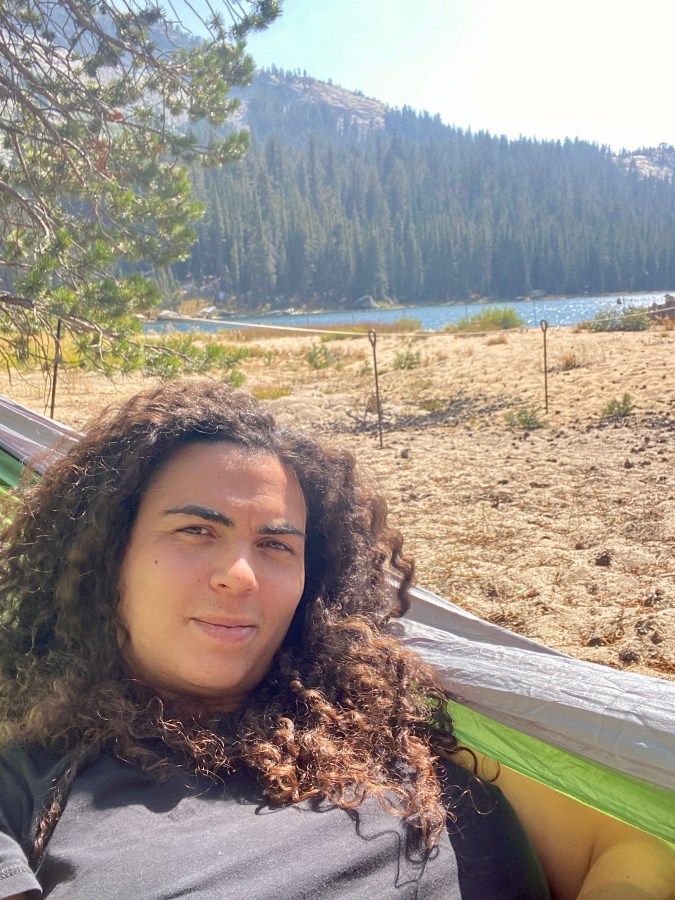 Abeni, a Black woman, lies in a hammock in front of what looks like a beach near a body of water also near a forest and mountains