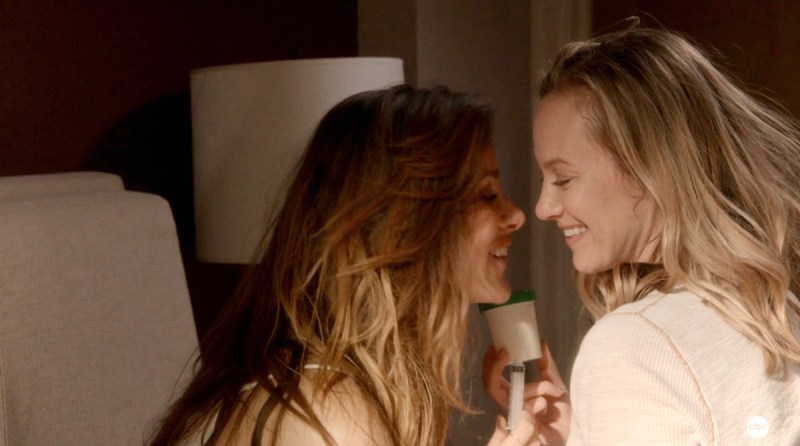 In a still from Station 19, Carina, a brunette with wavy hair, and Maya, a blonde with a blunt bob, sit together on their bed. Their faces are pressed close together, laughing. They hold up a cup of sperm and an insertion device not unlike a turkey baster.