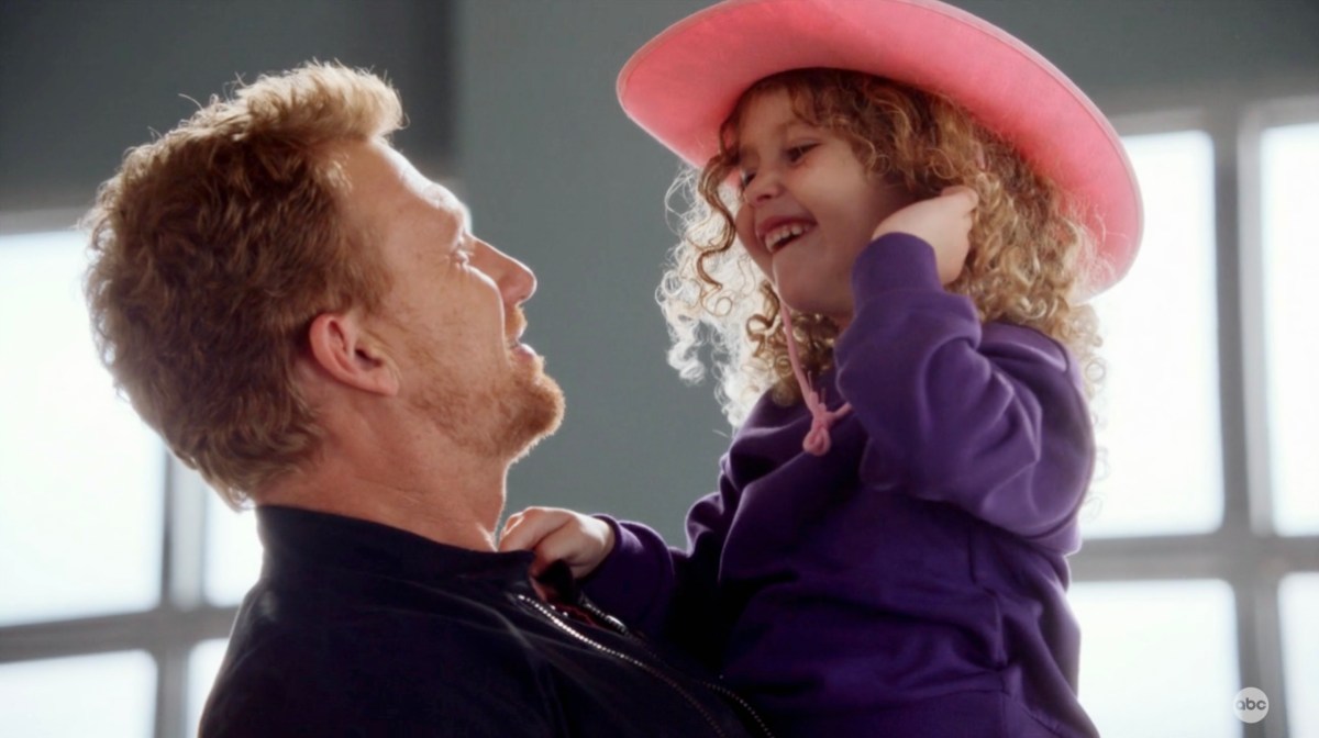 In a still from Grey's Anatomy, Owen Hunt, a middle age man with red hair, hold his daughter Leo, a four year old trans girl with curly red hair and a pink cowgirl hat, in his arms. Leo laughs at the camera.
