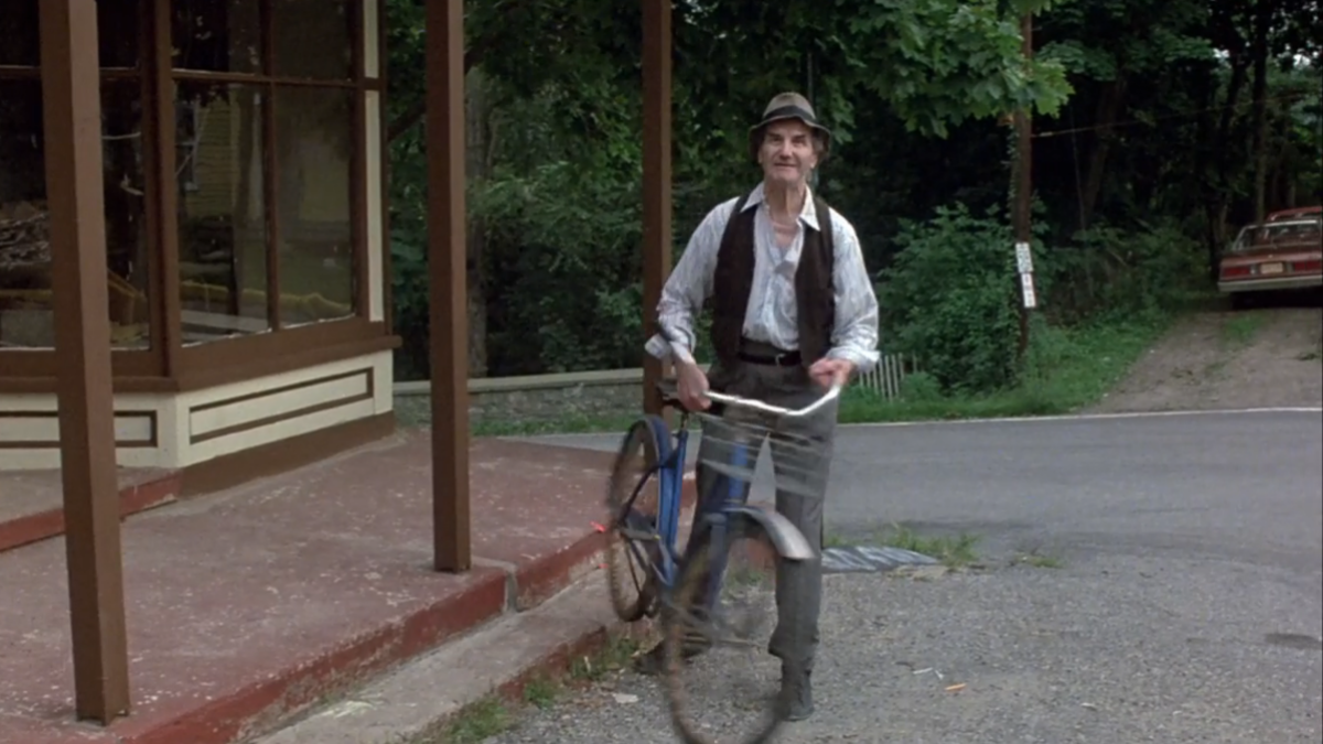 An old man wearing a vest, a buttondown shirt, slacks, and a fedora holds a bike in Friday the 13th