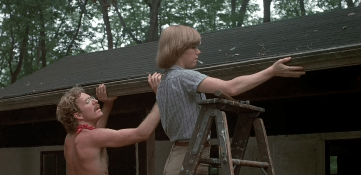 A shirtless white man and a white woman in khakis, a short sleeve buttondown, and a bowl cut work on a cabin roof in Friday the 13th