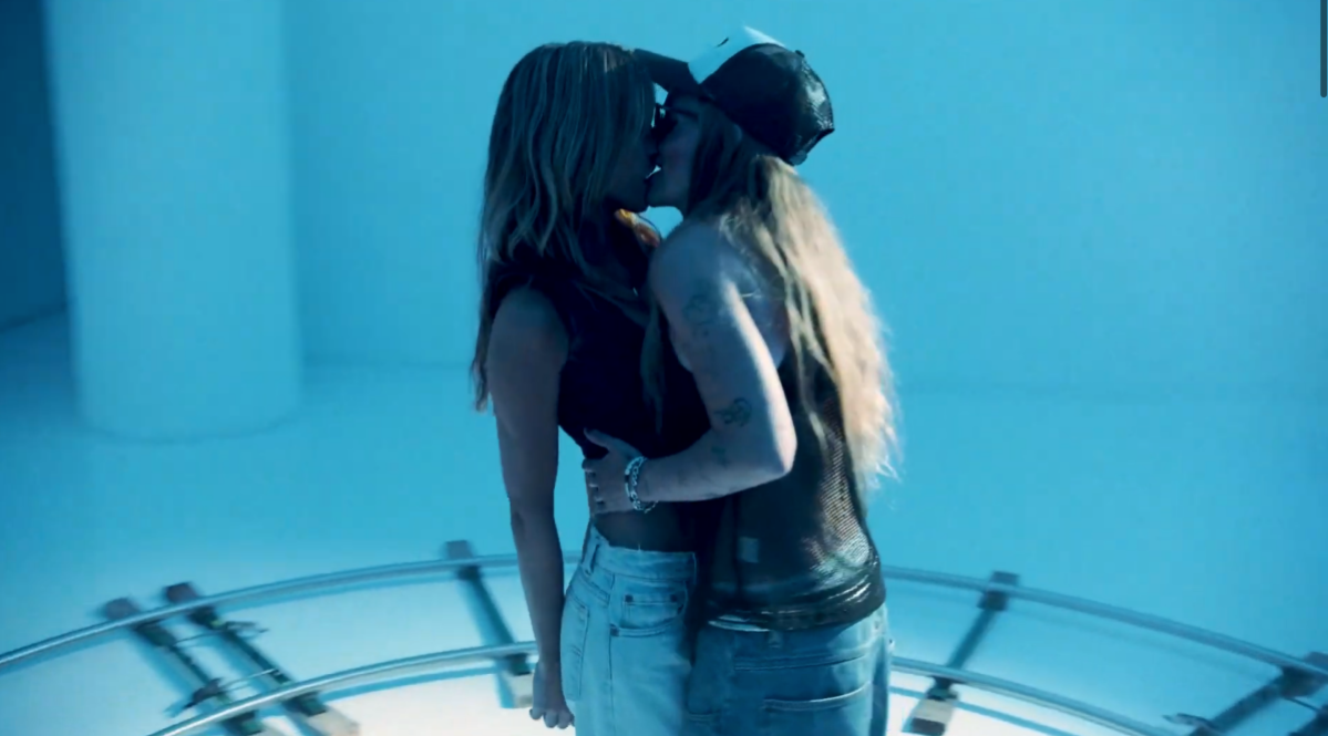In a still from "Get Me Outta Here," Chrishell Stause and G Flip are making out. Chrishell Stause is wearing jeans and a dark tank. G Flip is wearing jeans and a black mesh tank and a hat.