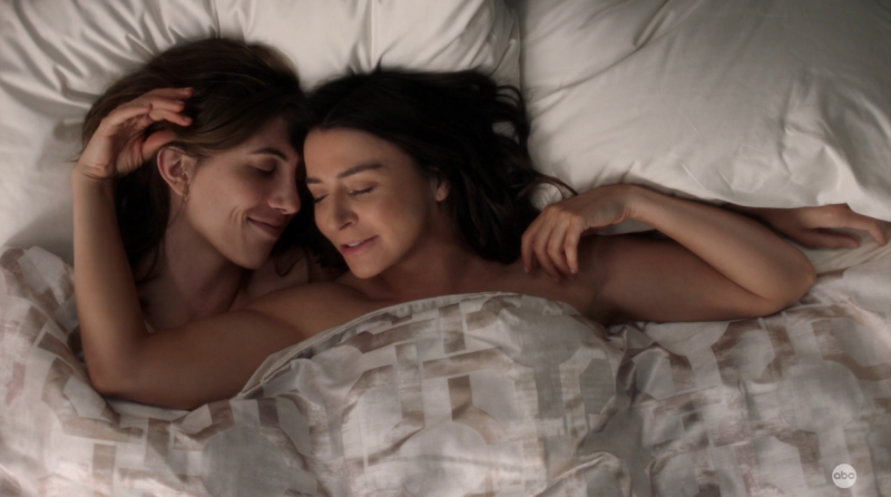 Kai and Amelia lay together in bed with a cover up to their necks and playing in each other's hair. They are smiling in bliss.