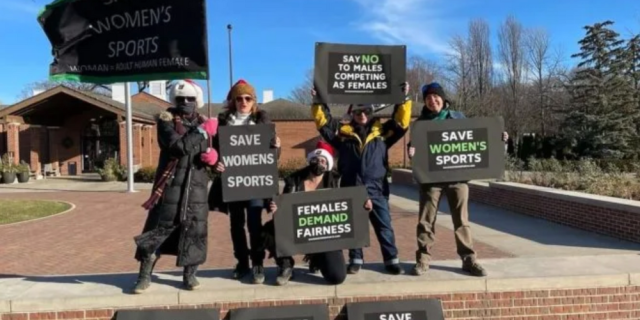 A group of protesters outside in sunlight holding signs that read "save women's sports" and "females demand fairness."