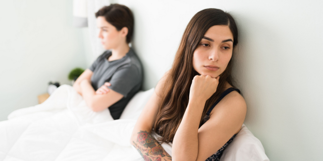 In a room with white walls, a woman with olive skin, long, brown hair and floral tattoos on her lower arm sits in a white bed with her chin in her hand. To her left, a woman with pale skin and short brown hair who is wearing a grey T-shirt sits with her arms crossed.