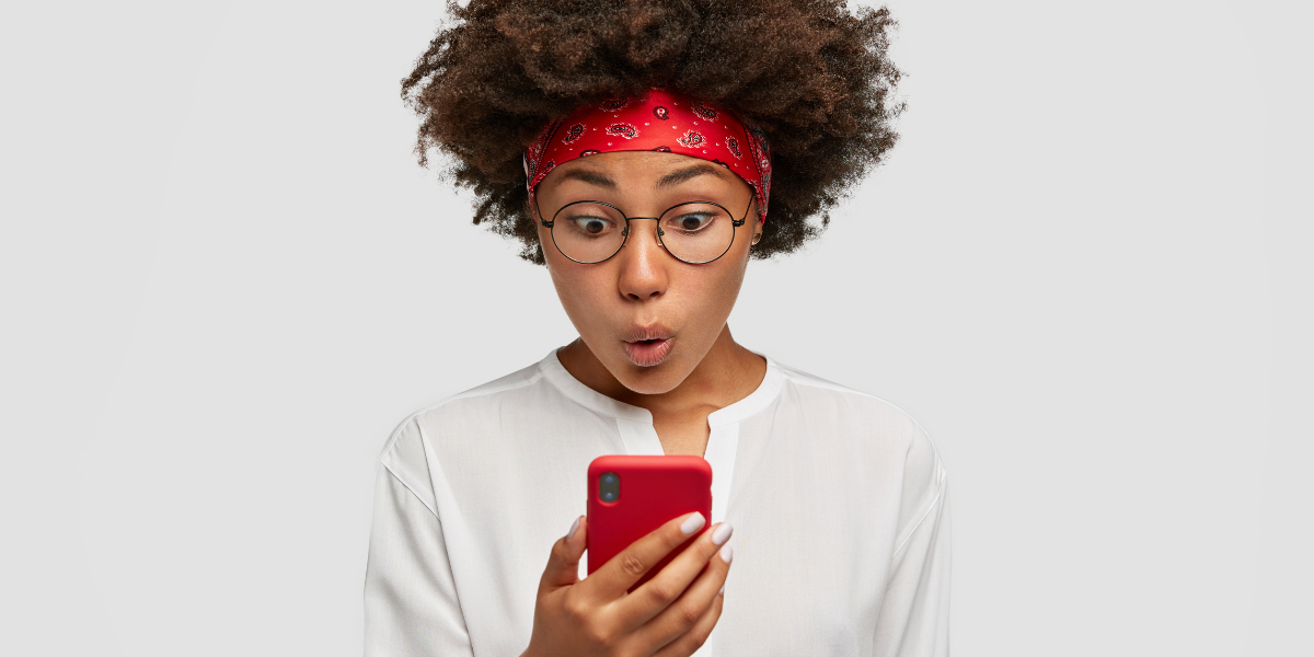 Against a gray background, a Black woman with natural hair who's wearing a red bandana around her head, wire-rimmed glassed, and a long-sleeve white shirt looks at her red phone with a surprised facial expression.