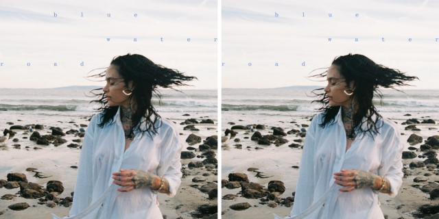 The album cover for Kehlani's "blue water road" features Kehlani on a stone-lined beach wearing a white buttondown, and their hair is blowing in the wind.