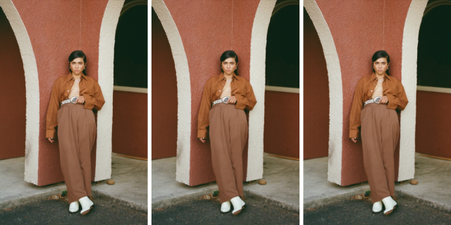 The musician Fabi Reyna has her hair slicked back and is leaning against an outdoor wall. They are wearing a brown bomber jacket that is open, wide legged brown trousers, a thick white belt, and white boots.