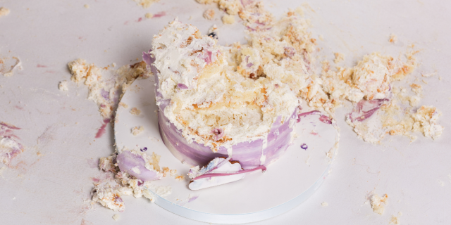 Against a white background, a yellow cake with pink and white frosting sits on a white plate. The top of the cake is fully smashed.