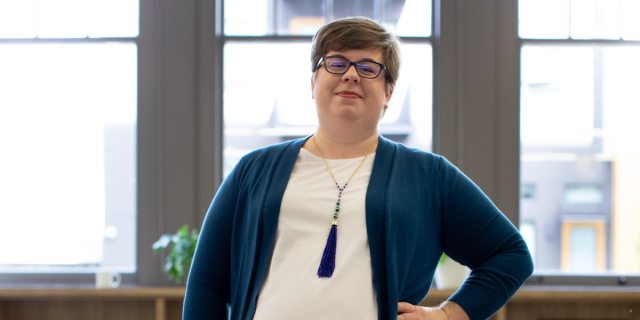 A fat person with a short haircut stands proudly with one hand on their hip in front of three bright windows, wearing a white t-shirt, blue cardigan, a fancy necklace, and thick black framed glasses.