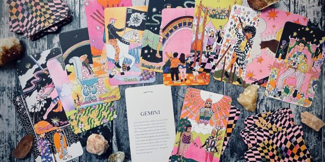 A collage of tarot cards on a grey wood table, the cards are in shades of Pink and spring warm hues. In the middle is a card that says Gemini in script font.