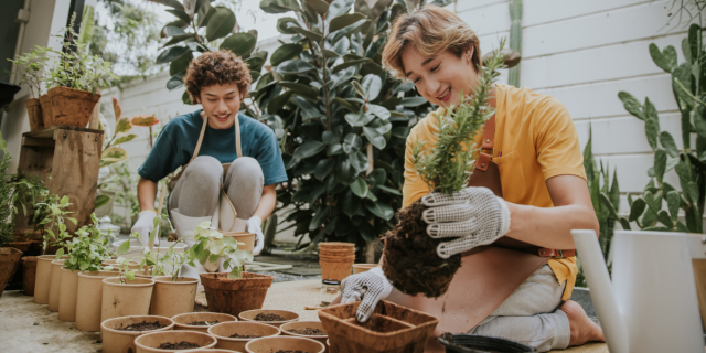 two genderqueer people re-pot plants and trees and vegetables with resuable pots for their home garden. They are smiling and look like they're having a delightful time.
