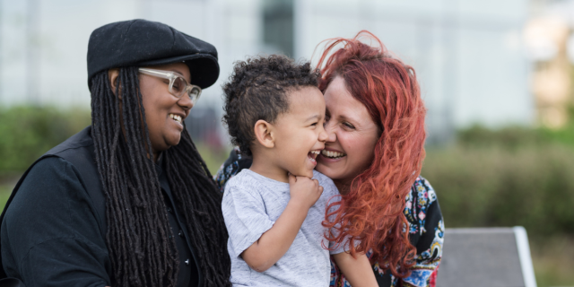 Two mothers hold and laugh with their child. The mother on the left is Black and wearing a cap, glasses and black shirt with hair in long locs. The mother on the right is white with dyed strawberry hair. The child is a mixed-race toddler with curly dark brown hair.