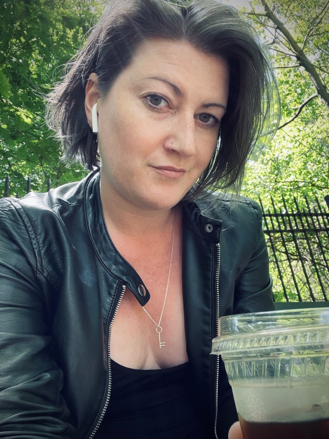 Meg, a white woman with gray hair, wears a black leather jacket and holds a cold beverage outdoors