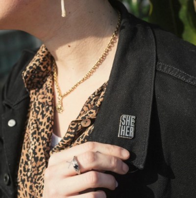 a gold and black she/her pin on the black jean jacket of the model, who has light skin and is wearing a leopard print shirt. the pin font is blocky and in all caps