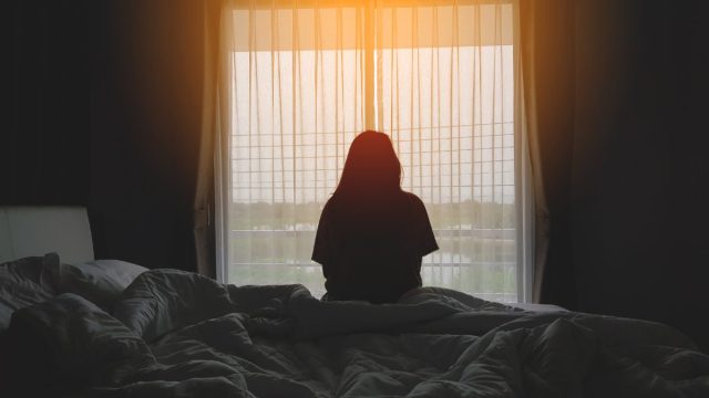 A person with long hair sitting on her bed, silhouetted by the window, facing the filmy transparent curtains. Outside, there is the hint of a sunrise.