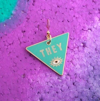an earring on an inverted triangle. the background is turquoise, and on top, is an eye and the word "they" in gold.