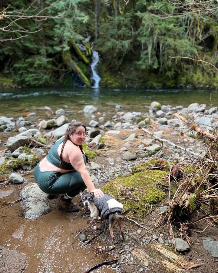 Vanessa, a white woman with her hair in long brown pigtail braids, poses with Zucchini by a stream, outdoors in the woods.