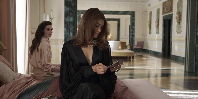 Anne Hathaway sits on a bed and stares lovingly at Zendaya over her shoulder