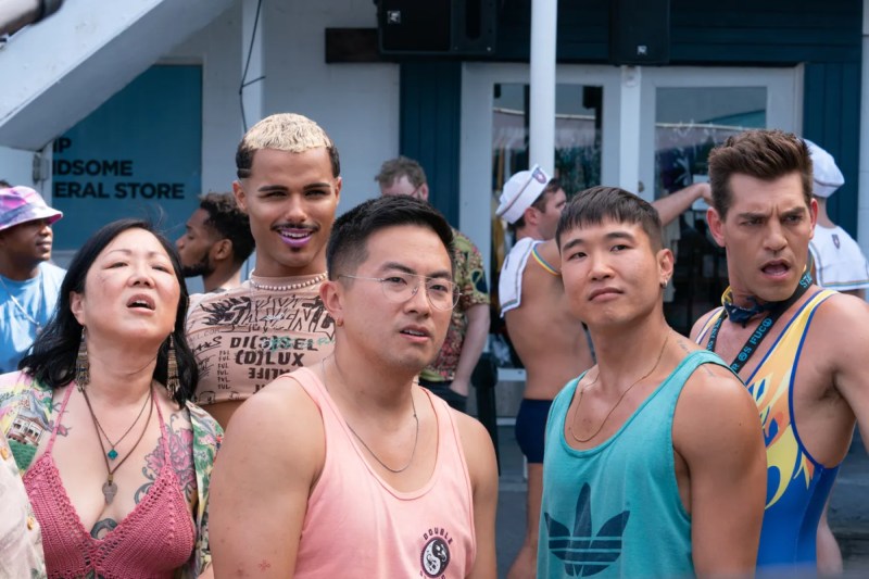 Margaret Cho stars in Fire Island, here she is in a red knit crop top showing off her tattoos while standing with her castmates (all of whom are gay men, also dressed for summer) on a boardwalk on Fire Island.