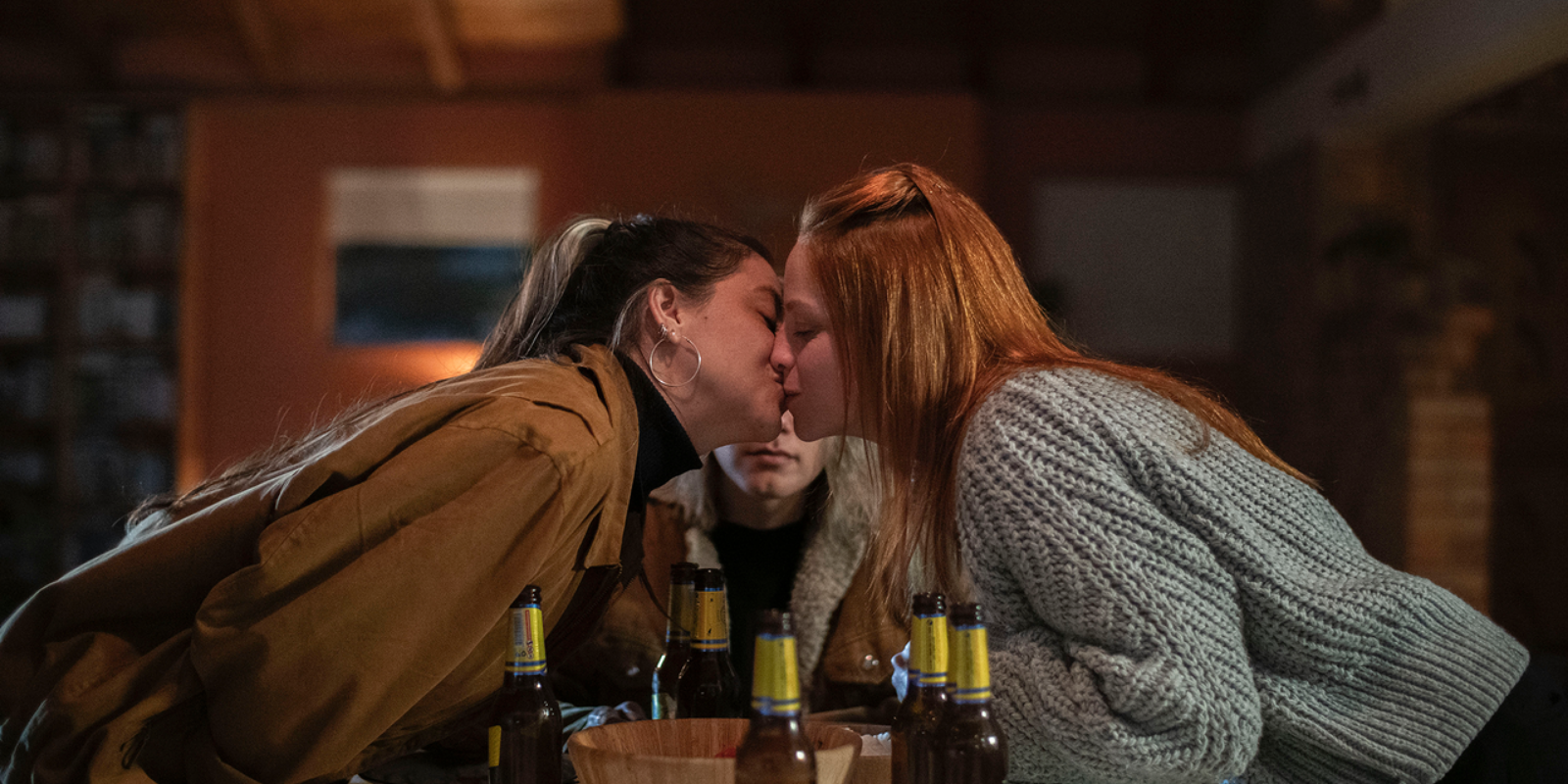 Two girls lean across the table and kiss while a third woman watches somewhat angrily.