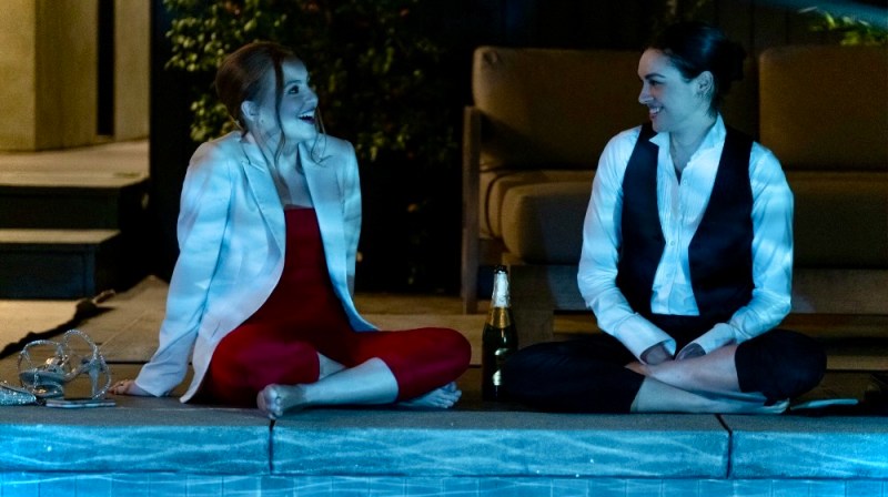 In Love Victor Season 3, Lake is in a red jumpsuit and white suit coat, sitting cross legged at a pool next to Lucy, who is in a cater waiter uniform that is unbuttoned.