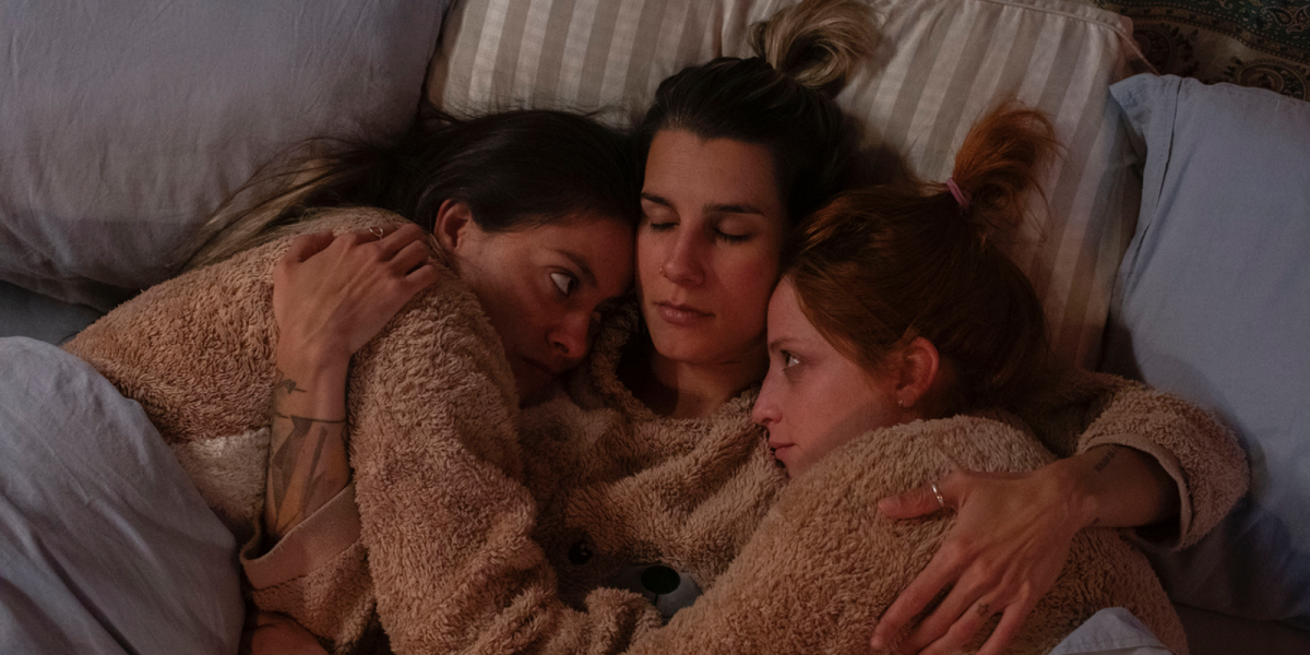 Three women cuddling in bed wearing furry onesies.