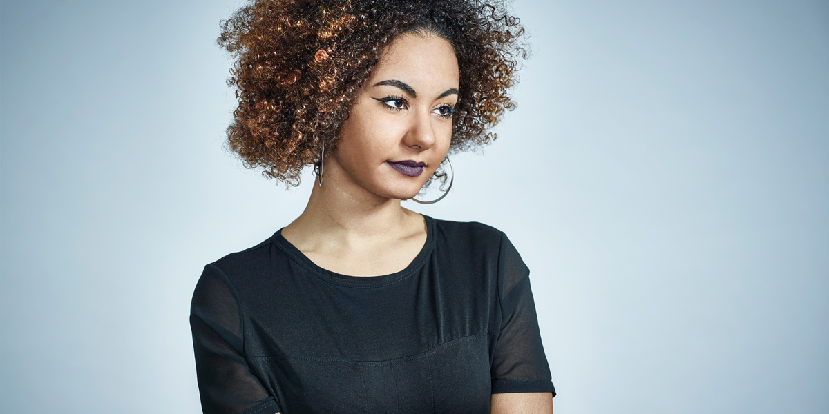 A light skinned black woman with a large brown afro has dark purple lipstick and a black t-shirt, she's looking off to the right of camera. She's in front of a blue background.
