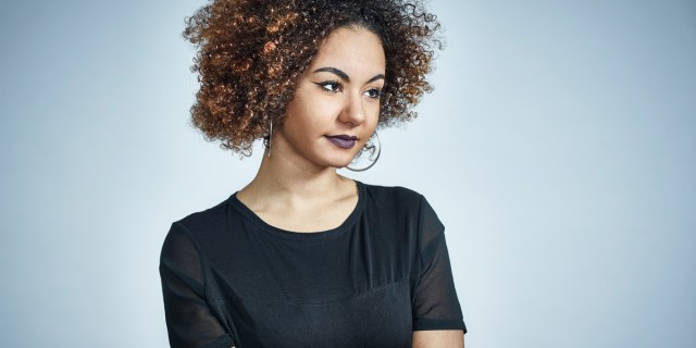 A light skinned black woman with a large brown afro has dark purple lipstick and a black t-shirt, she's looking off to the right of camera. She's in front of a blue background.