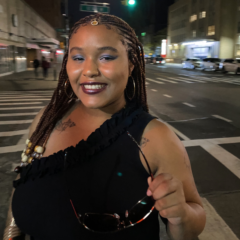 Amari is a Black person with long braids that have gold details outside in New York City at night