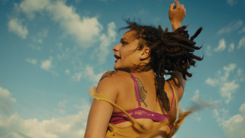 Sasha Lane as Star reaches her hand into the air hair and loose top blowing in the wind, clouds above her head