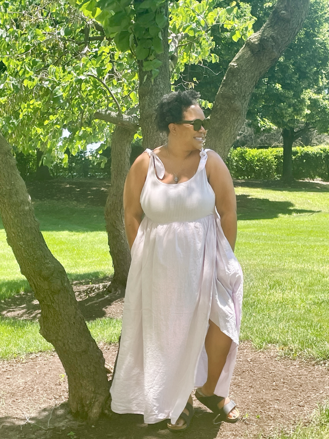 Carmen looks like an absolute goddess in this white or light pink full length sundress. She is a Black woman wearing sunglasses with medium-short curly black hair. Carmen is wearing Birkenstocks and smiling in front of a grove of trees.