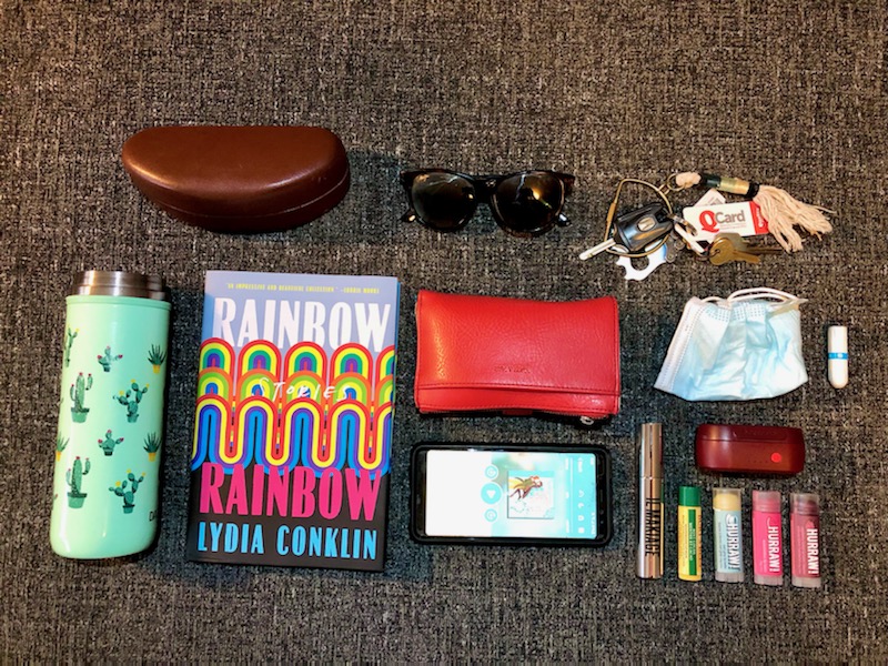 Casey's things are neatly arranged. There is a water bottle with cactuses on it, sunglasses and case, book, wallet in red, phone, multiple lip balms, headphone case, mask, tampon and keys.