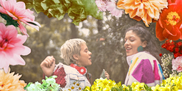 A woman with short blonde hair dances with headphones around her neck. She is wearing a cut off jean vest and a flannel underneath. She is dancing with another woman with brown hair in a ponytail who is wearing a windbreaker. They are surrounded by a floral border.