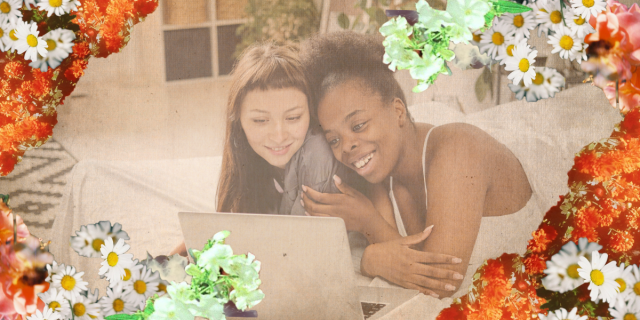 surrounded by flowers, a couple lies on a bed and looks on at a computer screen while smiling