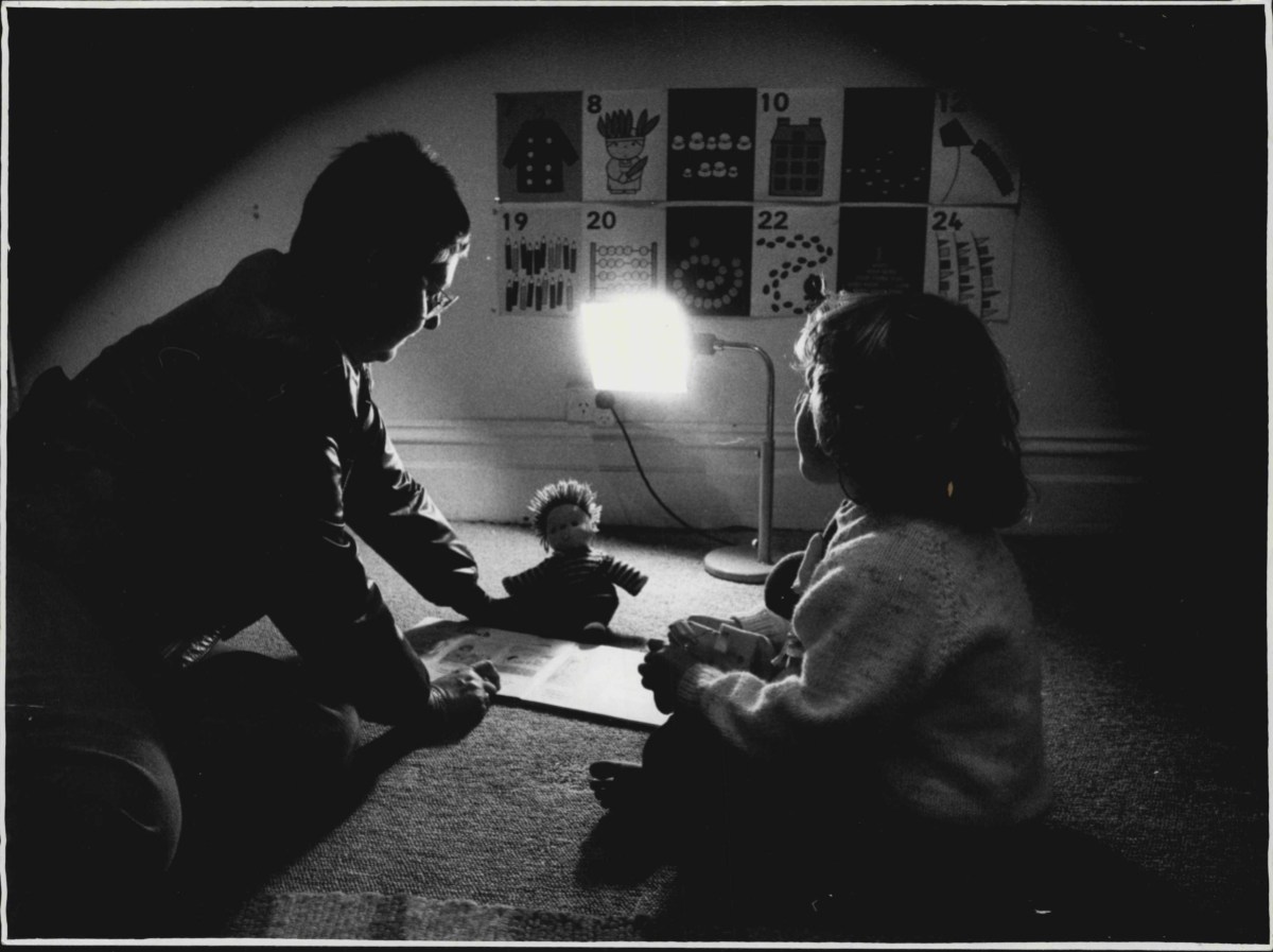 In a black and white photo that's dark except for a single light above a table, a mother and her daughter (both shadowed) sit across from one another with a small stuffed animal between them.
