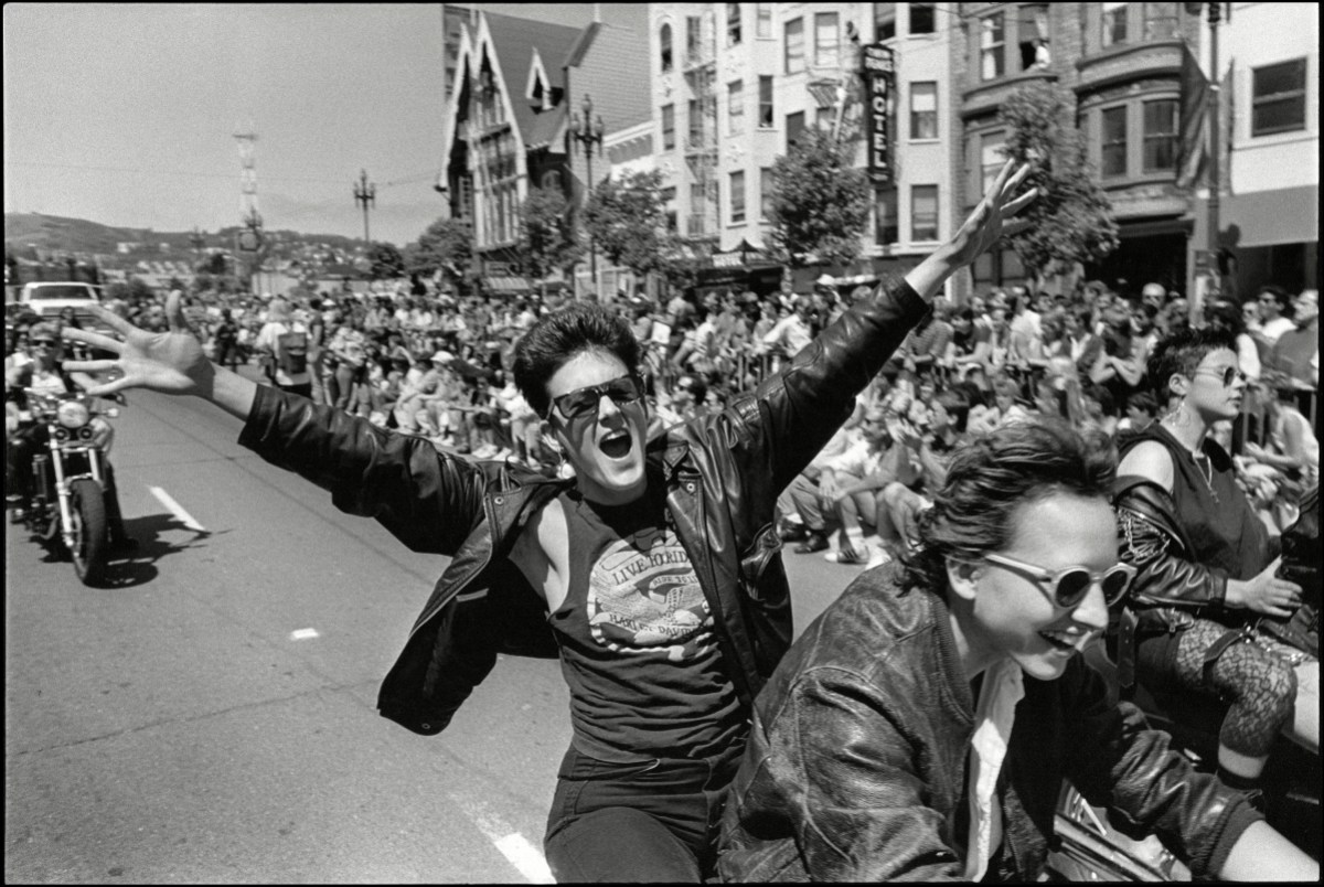 A woman, both her arms raised, rides on the back of a motorcycle in the 'Dykes on Bikes' group