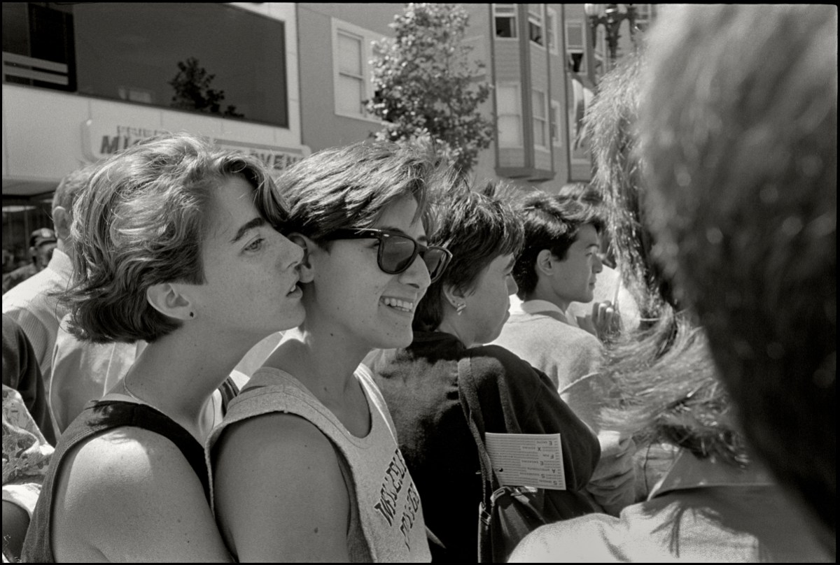 A black and white photo of two lesbians embracing, one is holding the other from behind. They both have short brunette hair.