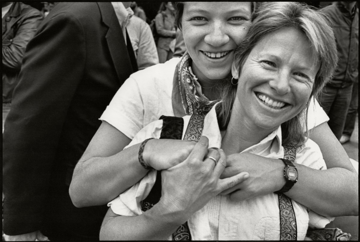 A close up of a white lesbian couple with short hair smiling in a black and white photo, one holds the other across her chest from behind.