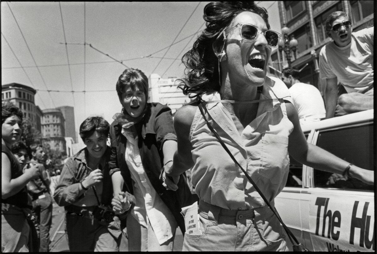 View of a group of women holding hands as they march, the woman in the front has her mouth open wide in laughter and is wearing sunglasses.