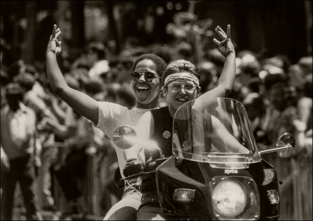 A black and a white woman share a motorcycle during dykes on bikes, the black woman has her hands up in celebration and is wearing sunglasses.