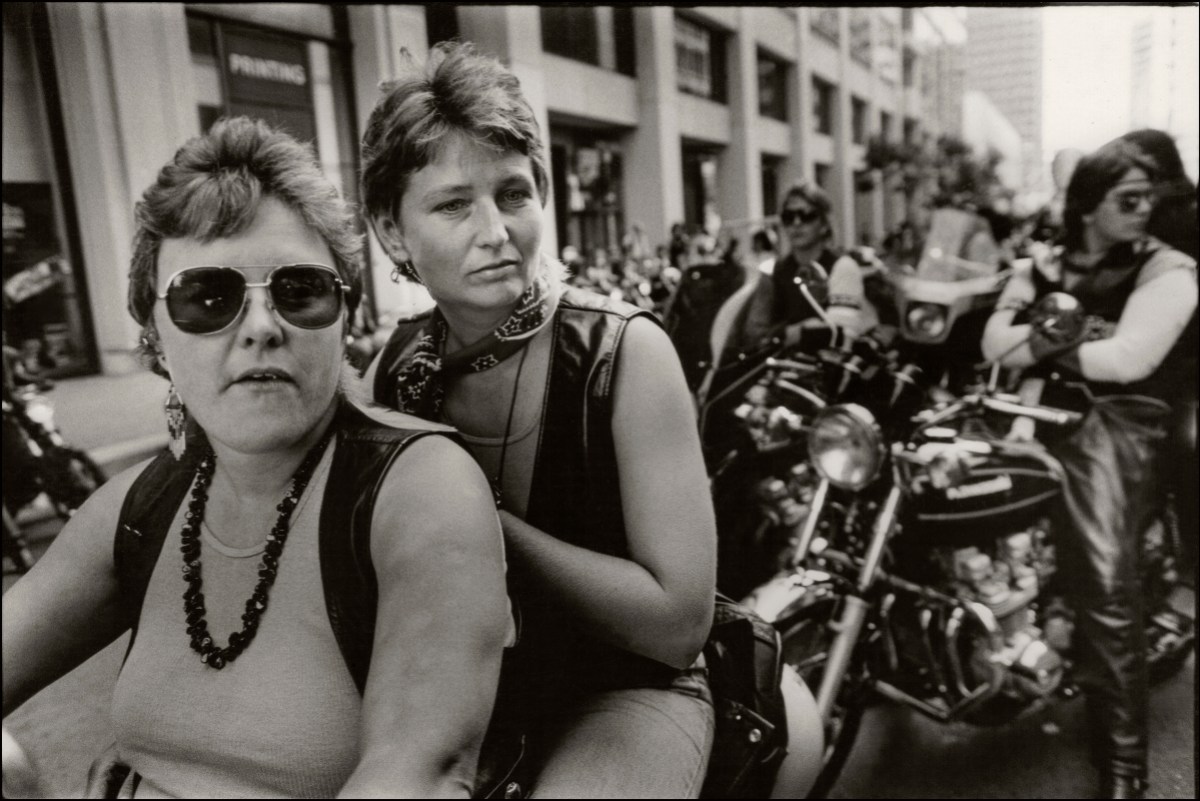 A close up portrait of two women at the dykes on bikes parade, they share a motorcycle and the first woman is fat wearing aviator sunglasses. The second woman is thin with a leather vest.