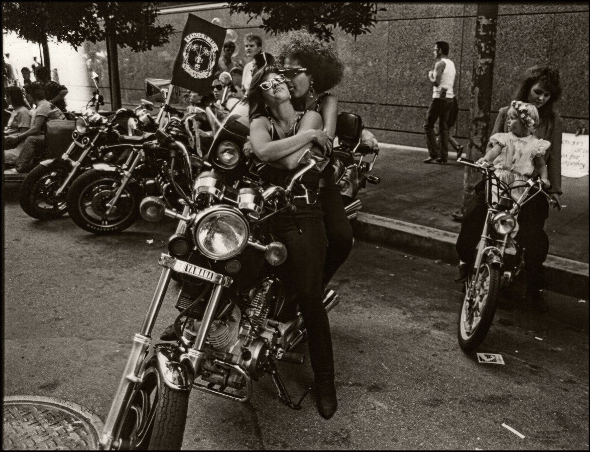 Two black women from Dykes on Bikes embrace, one is licking the ear of the other.