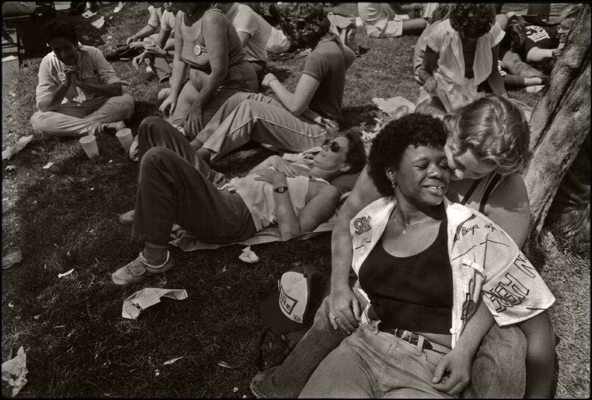 Among a crowd on the grass outside the Civic Center, two women share a smile together, one woman is black and lays across the lap of a woman who is white.