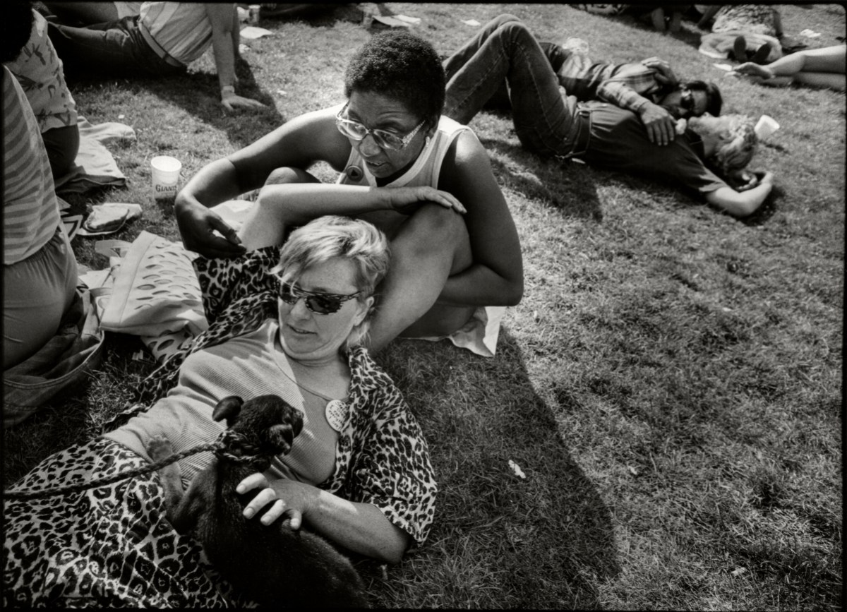 View of two women, one with her head against the shins of the other, as they sit, with a pet dog on the grass in a black and white photo. One woman is black and the woman propped up against her thighs is white.